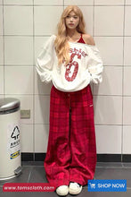Person wearing a white oversized sweatshirt with red text and red plaid pants in a tiled room.