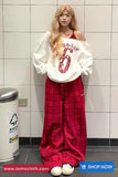 Person wearing a white oversized sweatshirt with red text and red plaid pants in a tiled room.