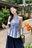 Woman in a blue top and dark skirt holding a drink, standing outdoors with greenery and a building in the background.