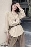 Woman wearing a beige pleated blouse and brown pants against a building wall.