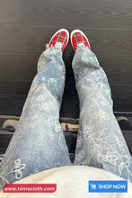 Person wearing red plaid shoes and light blue jeans with floral embroidery on a dark wooden floor.