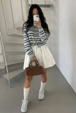 Person wearing a striped sweater, white skirt, white sneakers, and holding a brown handbag in a minimalistic room.