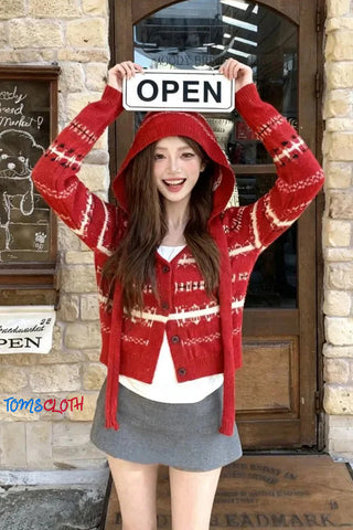 Person wearing a red sweater with white patterns, holding an 'OPEN' sign, standing in front of a door.