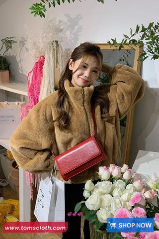 Woman in a brown coat holding a red bag with flowers and shopping bags in a room.