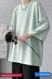 Person wearing a light green oversized shirt holding a black flower against a gray wall.