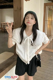 Woman in a white blouse and black shorts making a peace sign indoors.