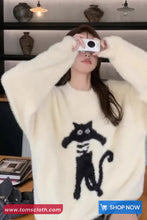 Person wearing a white sweater with a black cat design, holding a phone.