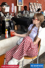 Person sitting at a cafe counter with a drink, wearing a white t-shirt and red checkered pants.