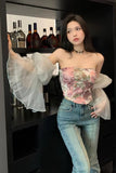 Woman wearing a floral strapless top and jeans in front of a shelf with bottles.