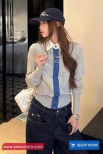 Woman wearing a gray cardigan with blue accents and a navy cap, standing indoors.