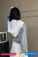 Person wearing a denim jacket with a white lace garment over it, standing against a plain wall.