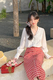 Woman in white blouse and red checkered skirt sitting outdoors with a red handbag and flowers.