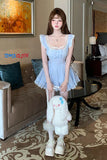 Woman in a light blue dress holding a plush toy in a living room with 'Toms Cloth' branding.
