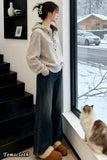 Person wearing a beige sweater and blue jeans standing by a window with a cat on a windowsill, snow outside.