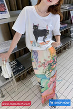 Woman wearing a white t-shirt with a black cat design and a colorful skirt in an indoor setting.