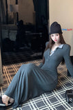 Woman in a long gray dress and black hat sitting on a patterned floor.
