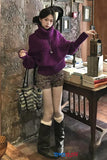 Woman in a purple sweater and black boots standing next to a table with books in an indoor setting.