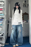 Person wearing a white floral top and blue jeans in a room with books and a bed.