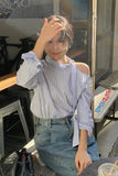Person sitting outdoors, wearing a striped shirt and jeans, with a blurred background.