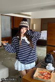 Woman in a striped hoodie and white skirt holding a pizza in a room with a bed and coffee table.