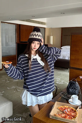 Woman in a striped hoodie and white skirt holding a pizza in a room with a bed and coffee table.