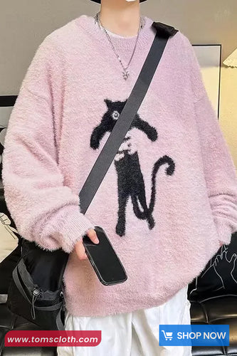 Person wearing a pink sweater with a black cat design, holding a phone, with a visible brand logo at the bottom.