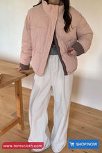 Person wearing a pink puffer jacket and white pants in a minimal indoor setting with 'tomscloth.com' branding.