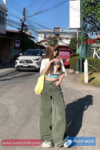 Woman standing on a street wearing a white crop top and green pants, with cars and buildings in the background.