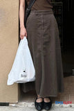 Person wearing a long brown skirt holding a white plastic bag, standing against a beige wall.