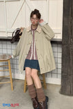 Woman wearing a beige coat over a plaid shirt and denim skirt, standing indoors.