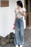 Woman wearing a floral top and blue jeans standing outdoors with a white wall and rocks in the background.