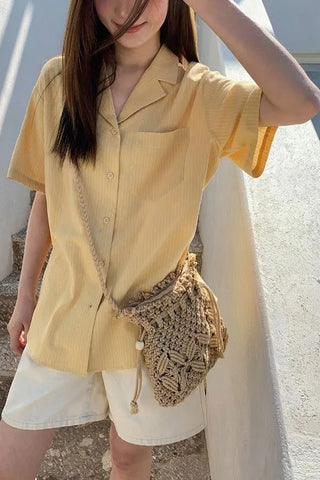 Person wearing a yellow shirt and white shorts with a woven bag, standing against a light-colored wall.
