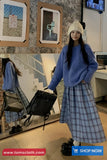 Woman wearing a blue sweater and plaid skirt standing in a room with framed pictures on the wall.