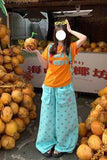 Person in an orange shirt and light blue pants standing among coconuts with a blurred face.