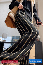 Person wearing a long, dark-colored coat with striped pattern, holding a brown bag indoors.