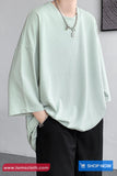 Person wearing a light green oversized shirt and black pants against a gray background with 'TomsCloth.com' branding.