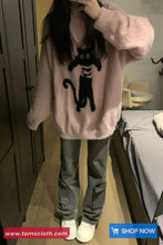 Person wearing a pink fluffy sweater with a black cat design, standing in a room.