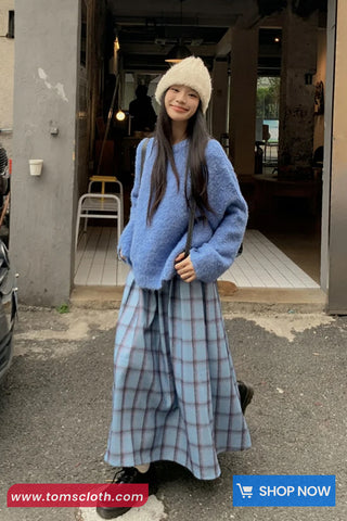 Woman wearing a blue sweater and plaid skirt standing outside a building.