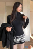 Woman wearing a black turtleneck and patterned skirt holding a black coat indoors.