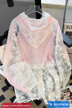 Pink lace top with sheer overlay on a hanger against a blurred indoor background.