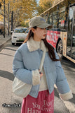 Woman in a blue puffer jacket with white fur trim standing on a street with a bus and trees in the background.