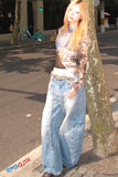 Woman in a sequined top and wide-leg jeans standing on a sidewalk next to a tree.