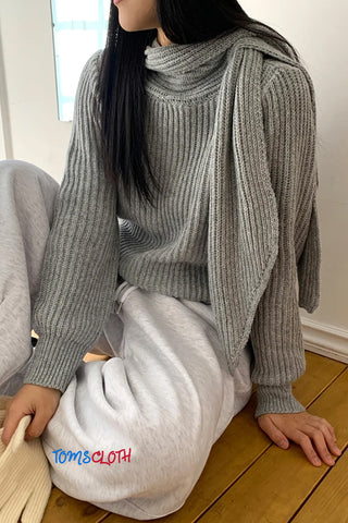 Person wearing a gray knit sweater and white pants sitting on a wooden floor.