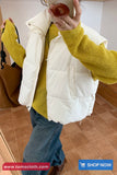 Person wearing a white puffer vest over a yellow sweater with a brown bag.