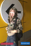 Person wearing a camouflage hoodie and blue jeans standing in front of a yellow wall with a large black and white logo.