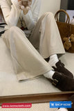 Person wearing beige pants and brown shoes sitting on a bed with a teddy bear and handbag in the background.