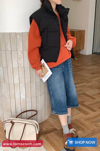 Person wearing an orange sweater, black vest, and blue jeans standing indoors.